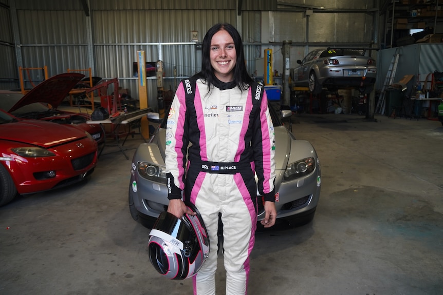 A young woman wearing motorsport gear and standing in front of a car.