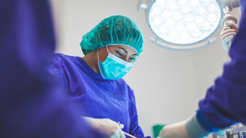 A woman performs surgery in scrubs and facemask
