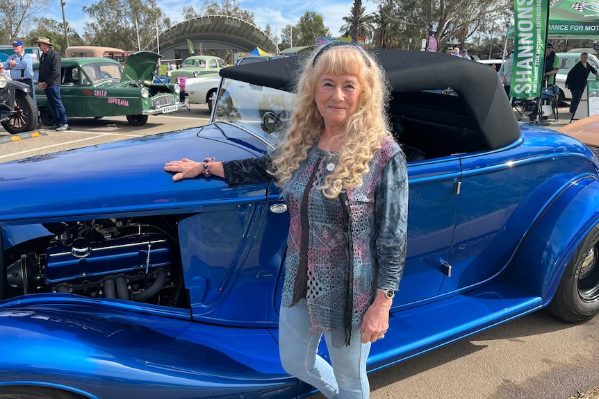 Woman stands with hand on the bonnet of a classic electric blue car