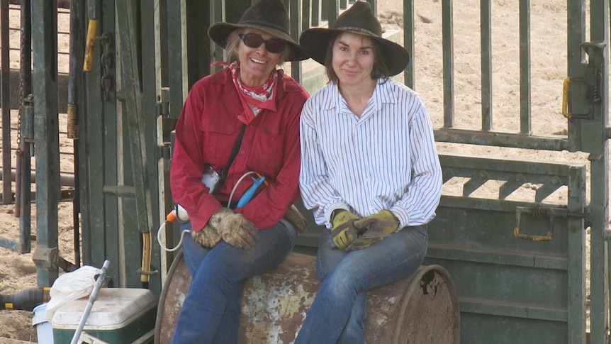 Two women sitting side by side on a rusty oil drum lying on its side.