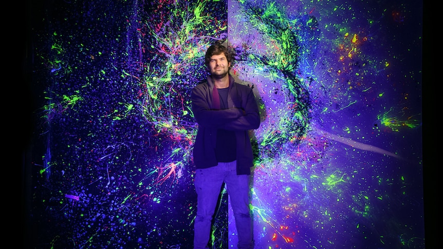 Man smiling against a large painting with luminescent colours.