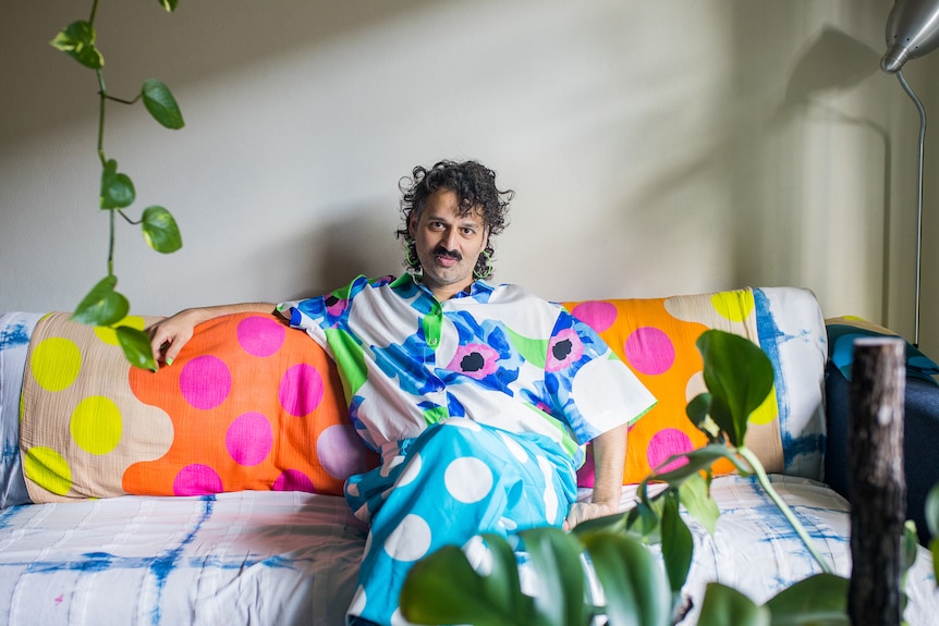 A Maltese Australian man with grey curly hair wears a vibrant floral shirt and neon green earrings, seated on a colourful couch