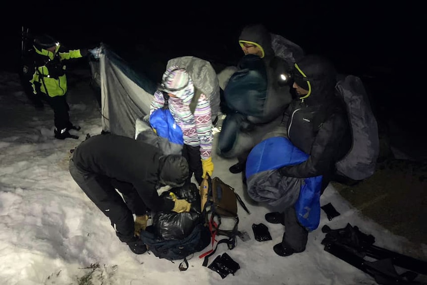 A group of people holding sleeping bags huddle around in the snow.