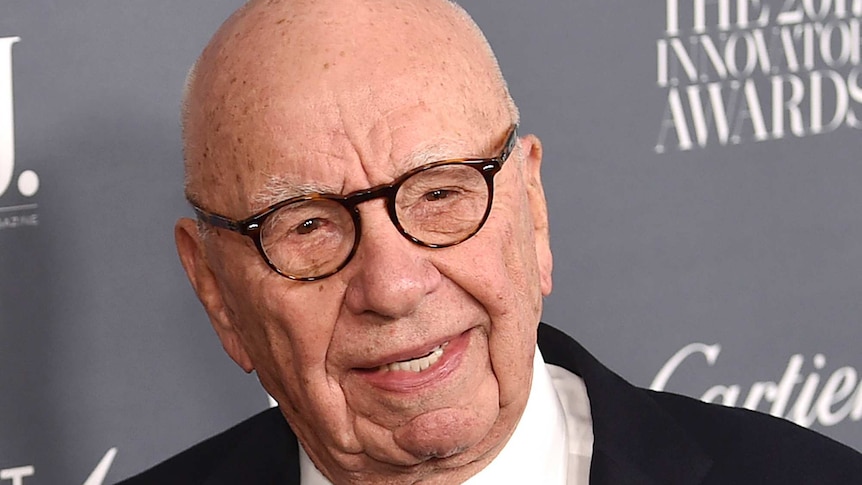 Tight shot of Rupert Murdoch standing in front of an advertising placard.