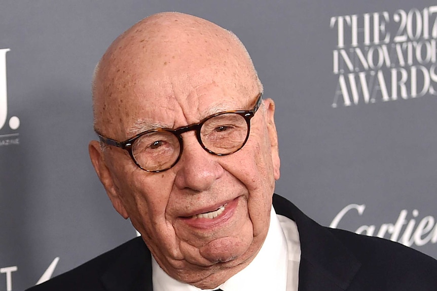 Tight shot of Rupert Murdoch standing in front of an advertising placard.