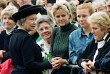 Queen Elizabeth, in a black coat and hat, wears a worried expression on her face while greeting a crowd of women