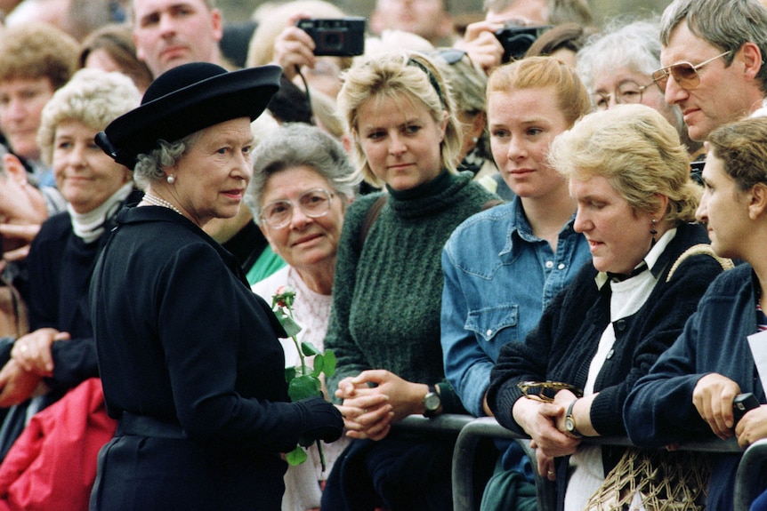 Queen Elizabeth, in a black coat and hat, wears a worried expression on her face while greeting a crowd of women