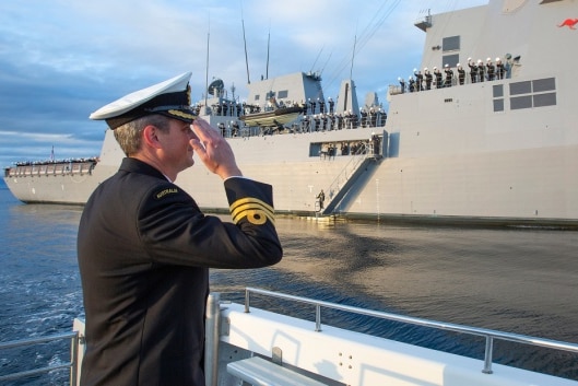 A naval officer with back turned to camera salutes a warship carrying seaman in the distance