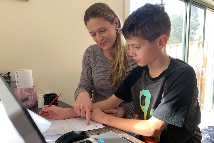 Suzanne sits next to her son at a desk with a laptop and a worksheet. She's pointing at the worksheet, he's holding a pencil.