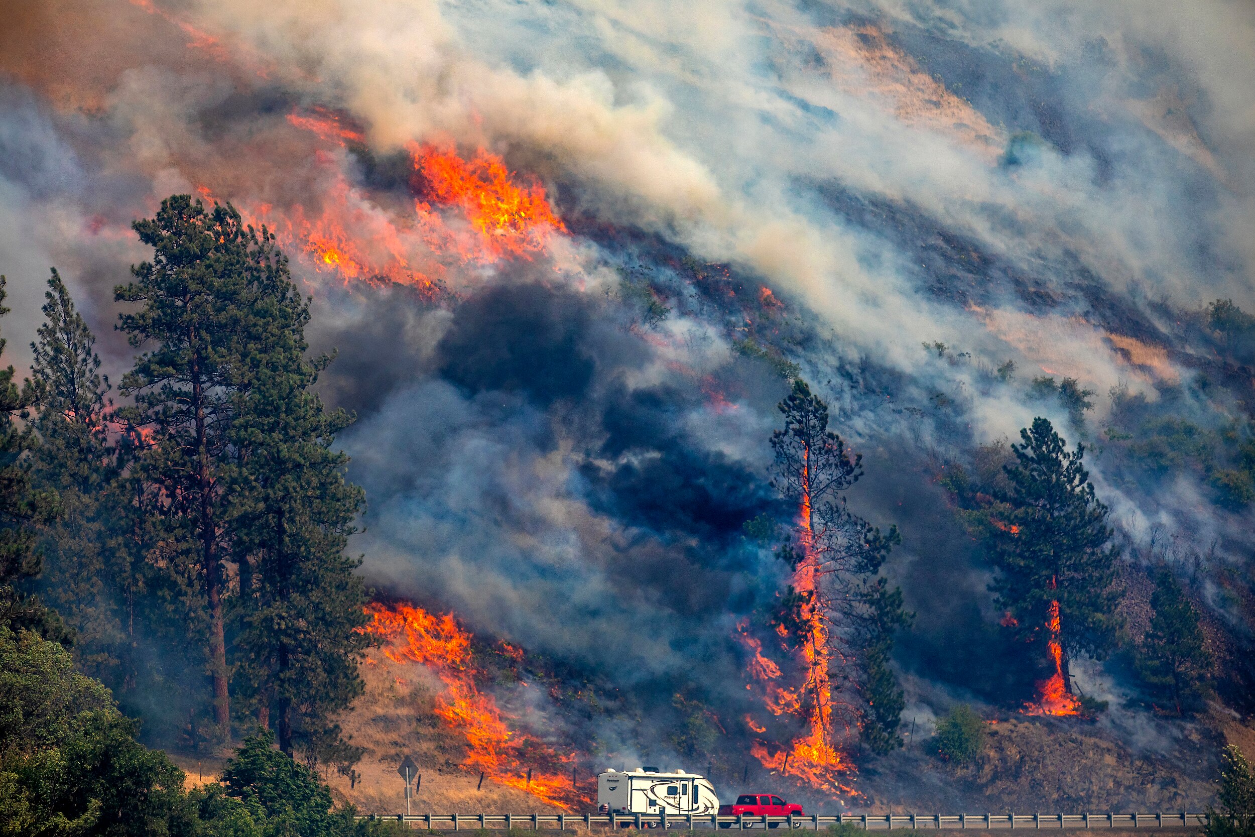A red car pulls a white caravan past a burning forest.