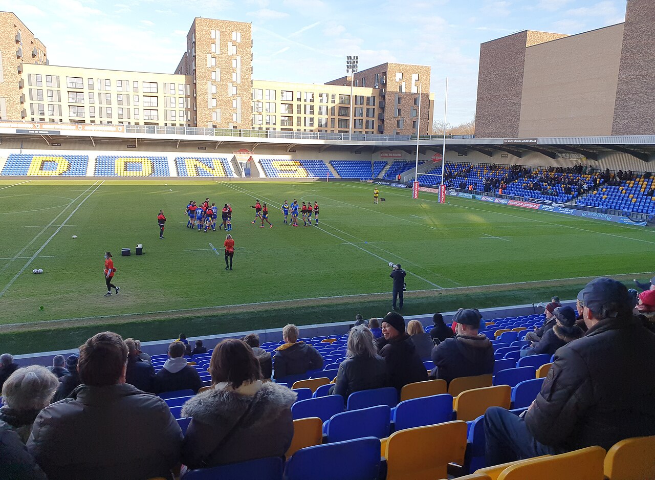 Players warm up on a rugby pitch, as seen from the stands.