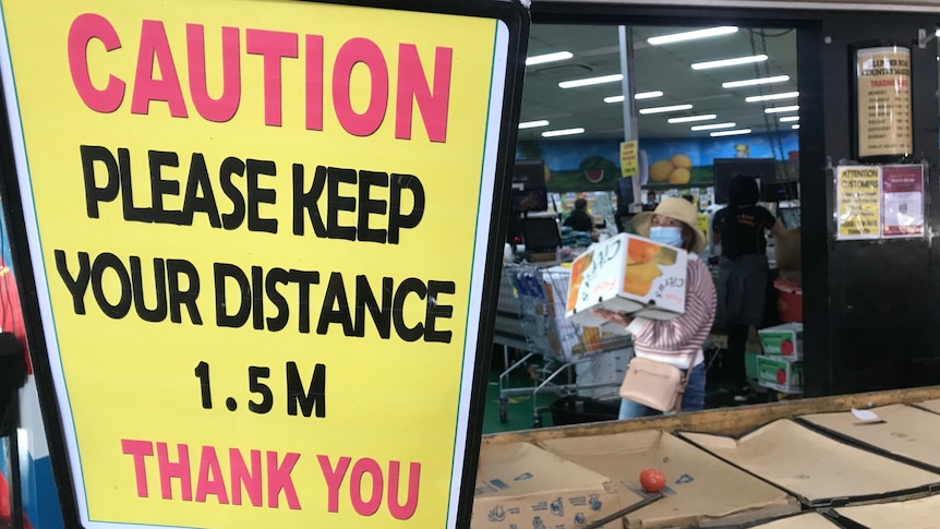 A masked woman carries a large box with a sign in the foreground asking people to stay 1.5m apart.