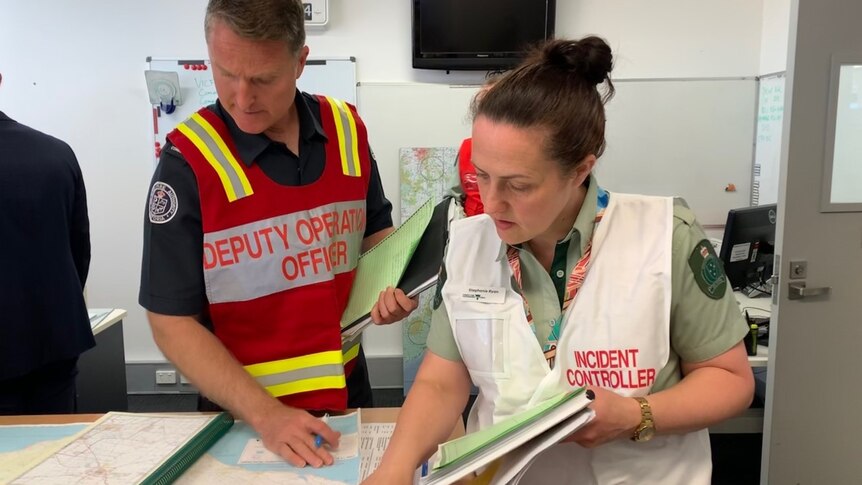 A woman in a green uniform and a white safety vest which says 'incident controller' looks at a map with a male police officer.