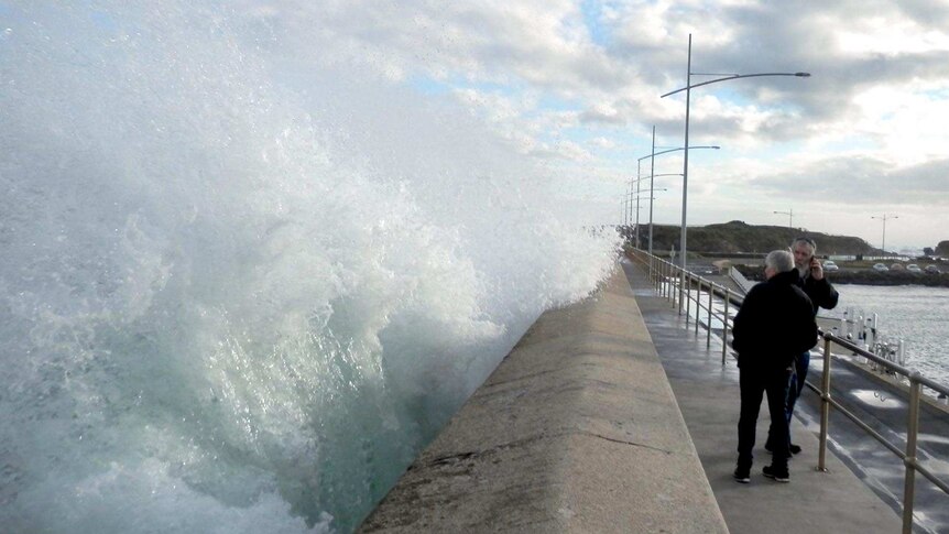 A wave smashes against the Warrnambool Breakwater right next to two people.