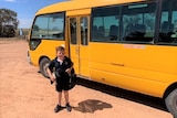 A young boy in black uniform and a backpack stands next to his yellow school bus parked on the red dirt and blue sky.