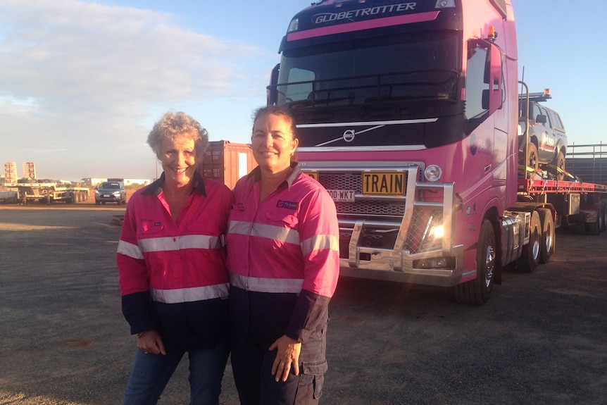 Two women in foreground smiling, wearing pink high vis work wear with large pink road train vehicle in background
