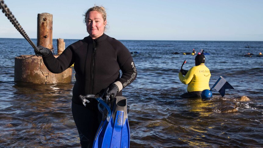 A blonde woman in a wet suit holding flippers stand in knee deep water, in the background people are snorkelling.