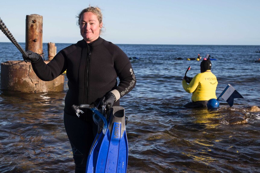 A blonde woman in a wet suit holding flippers stand in knee deep water, in the background people are snorkelling.