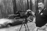 Black and white photo of Drought standing with camera with burnt out cars in background.