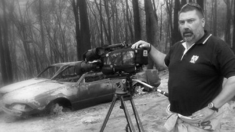 Black and white photo of Drought standing with camera with burnt out cars in background.