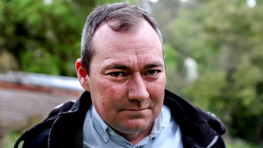 Man wearing blue collared shirt and black hooded jacket stares with green trees in background