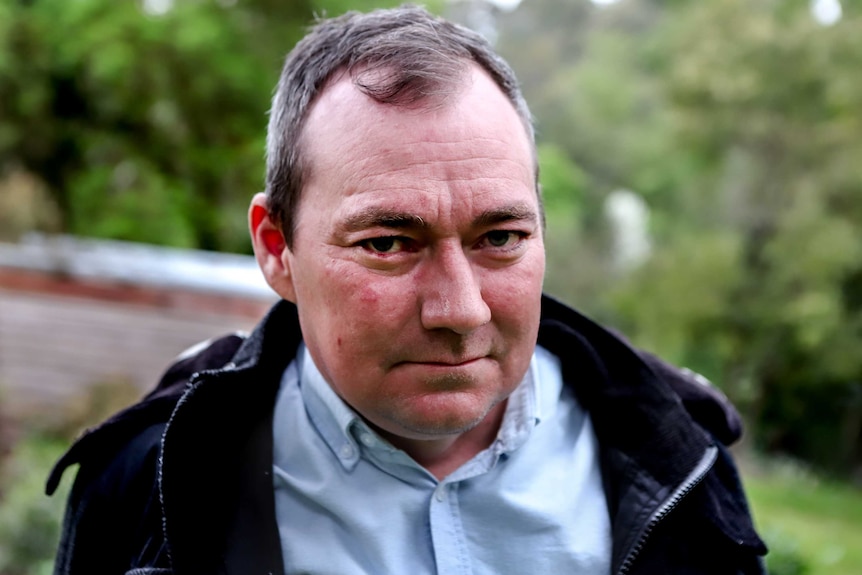 Man wearing blue collared shirt and black hooded jacket stares with green trees in background