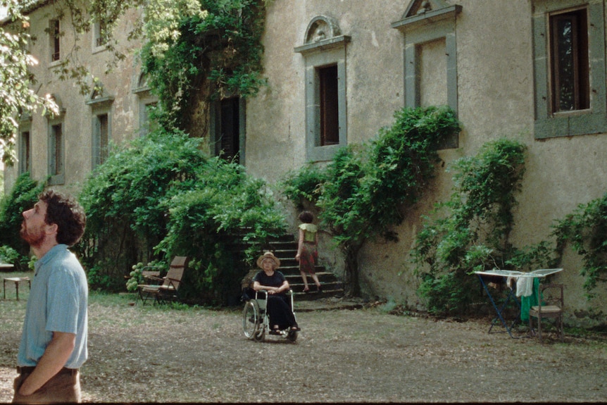 Exterior shot of an ageing Tuscan villa, with an older woman seated outside in a wheelchair.