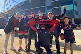 A small group of people in Melbourne Demons colours smile happily outside the MCG on a sunny day.