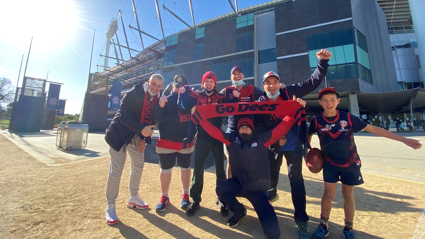 A small group of people in Melbourne Demons colours smile happily outside the MCG on a sunny day.