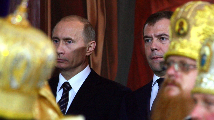 Vladimir Putin and Dmitry Medvedev, wearing black suits, are visible between several clerics wearing ornate head gold pieces