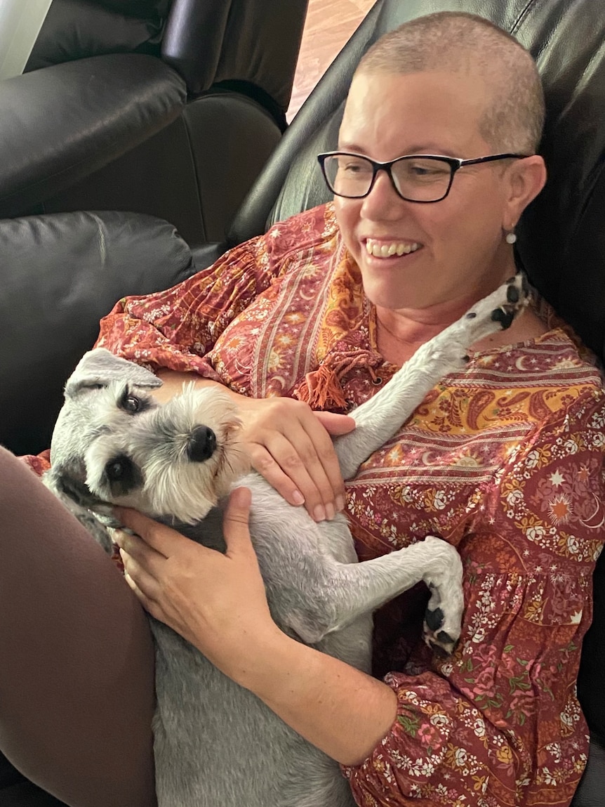 A woman with a shaved head cuddling a dog and laughing.
