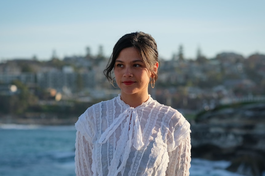 A young woman in a white shirt looks into the distance with a backdrop of Bondi beach behind her