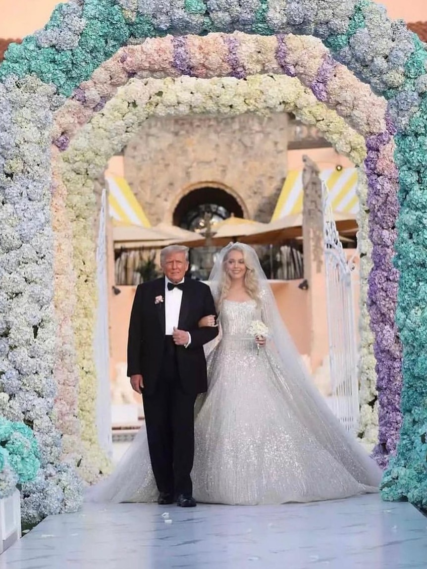 Donald Trump, wearing a suit, walks with his daughter Ivanka, in a jewelled wedding gown, through giant hydrangea arches