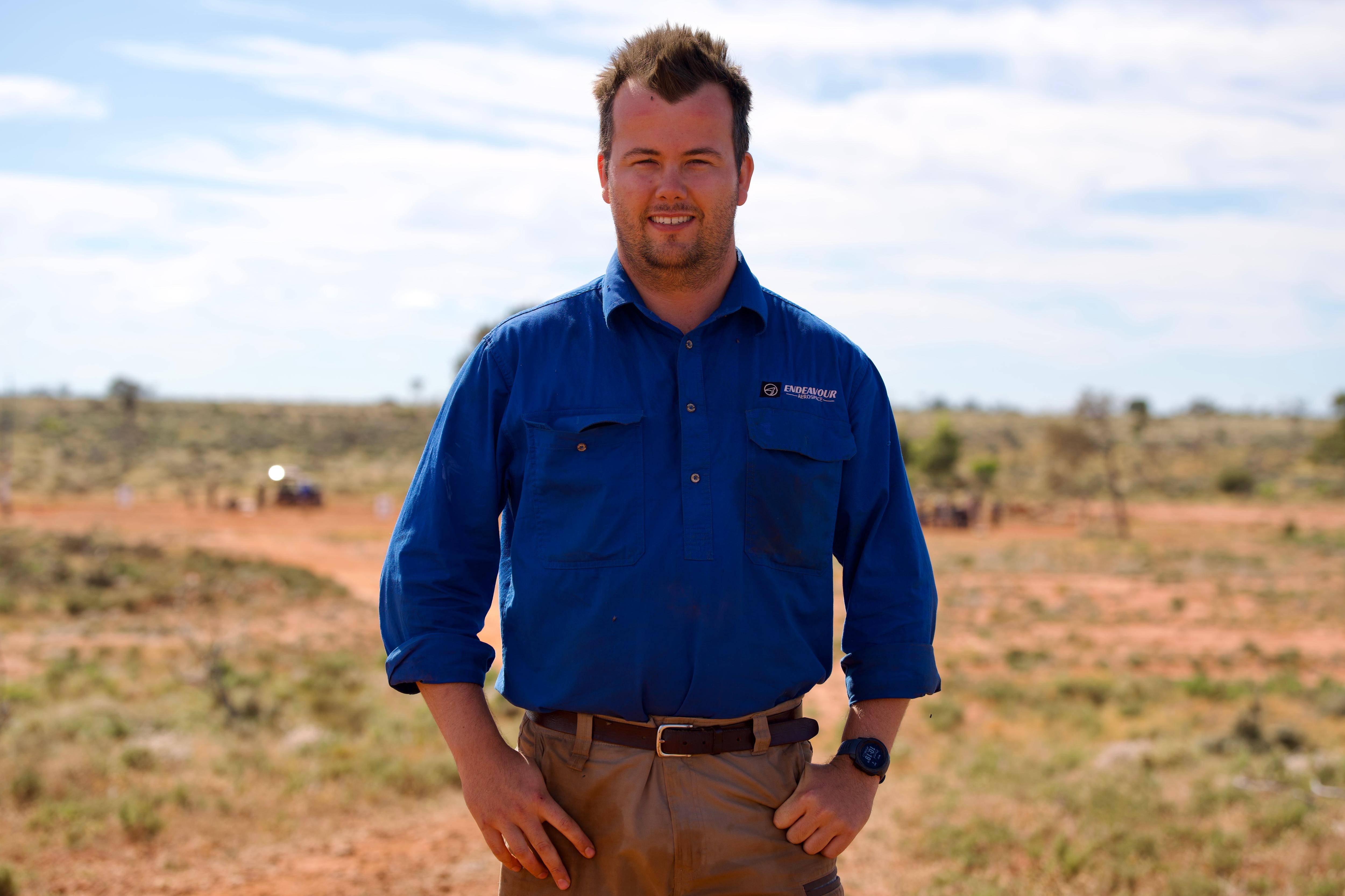 A smiling, dark-haired  man in a work shirt stands on an outback property.