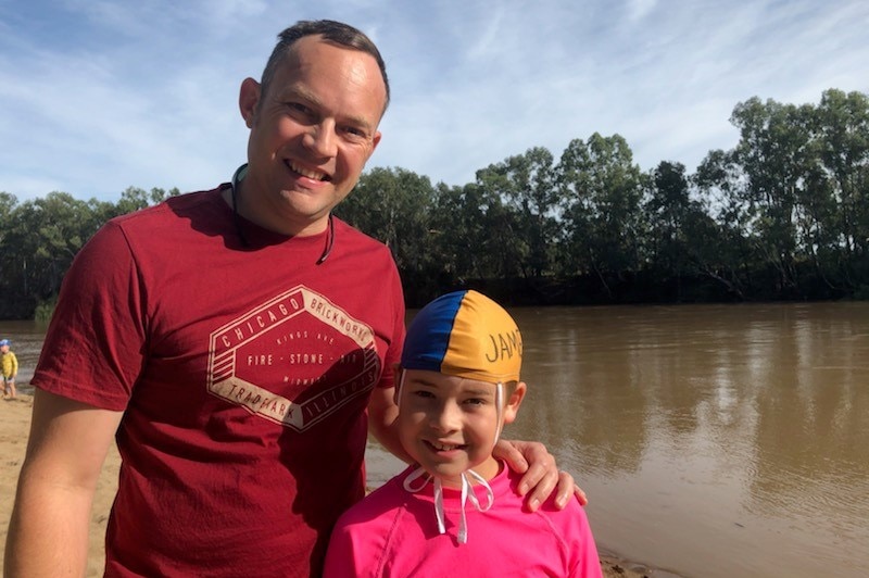 A man in maroon shirt with his arm around a boy in pink standing in front of a river.