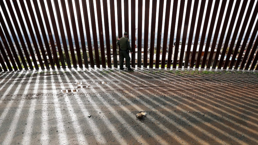 A border agent touches a wall of vertical steel slats lining a mud road creating dramatic parallel shadows on the road.