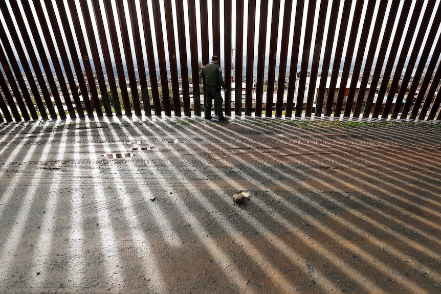 A border agent touches a wall of vertical steel slats lining a mud road creating dramatic parallel shadows on the road.