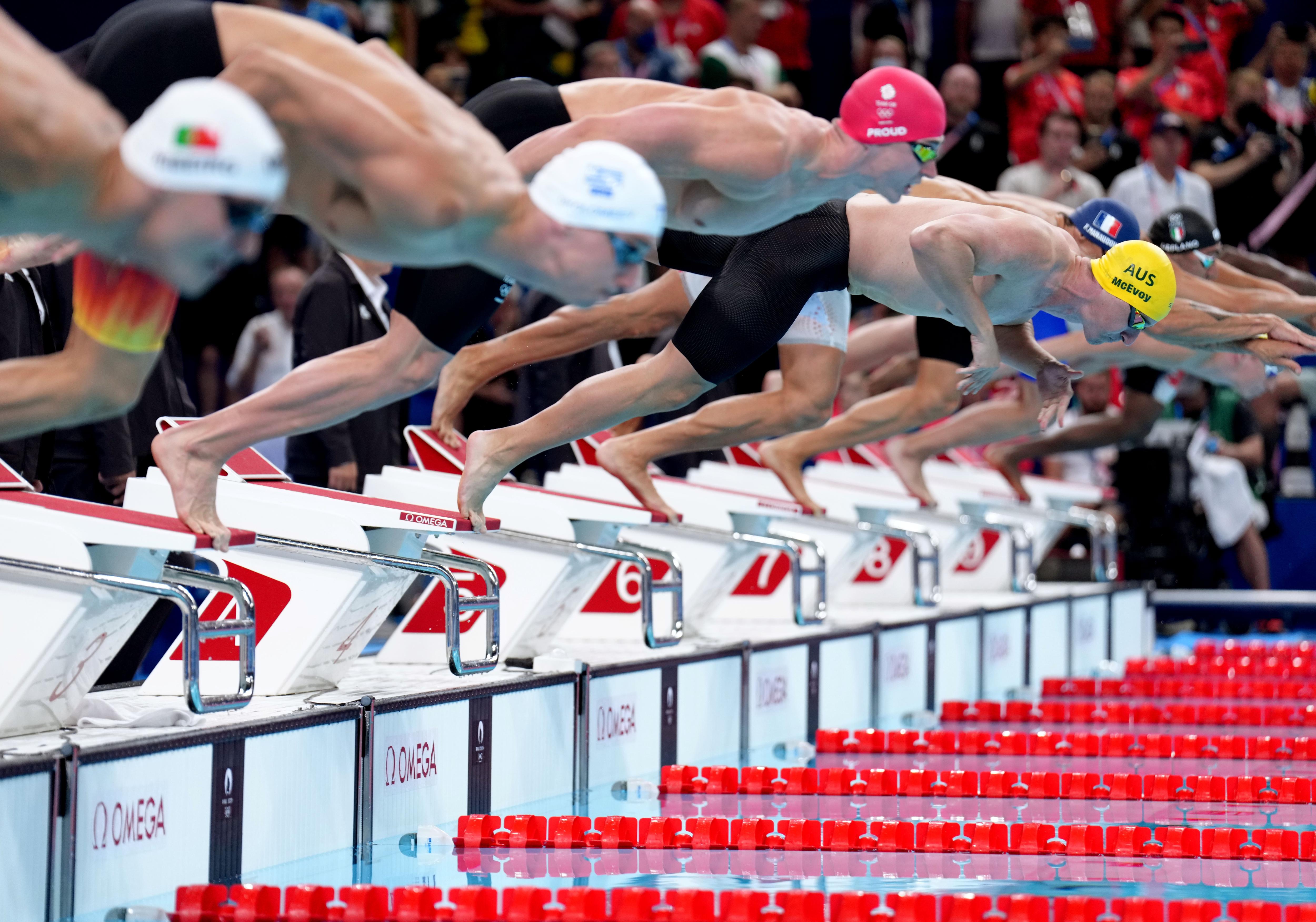 Male swimmers dive into the pool, wear caps, two whites, red and yellow, most in black, white, shorts, pink lane dividers.
