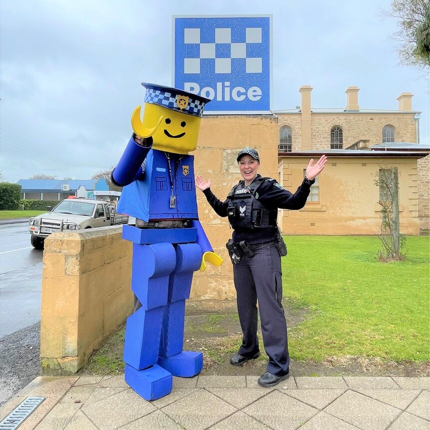A man wearing a Lego police officer costume with a female police officer outside a police station.