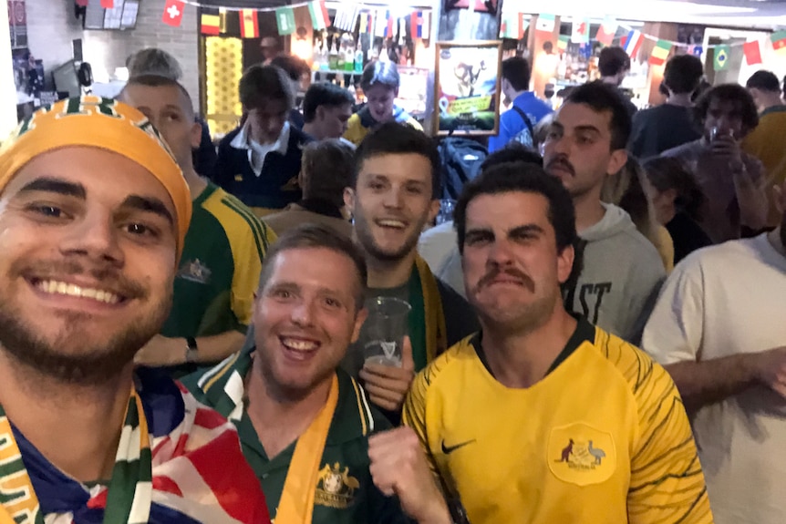 Grupė besišypsančių jaunų vyrų, apsirengusių australiškomis futbolo juostelėmis, stovi miesto gatvėje ir triumfuoja kumščiais.