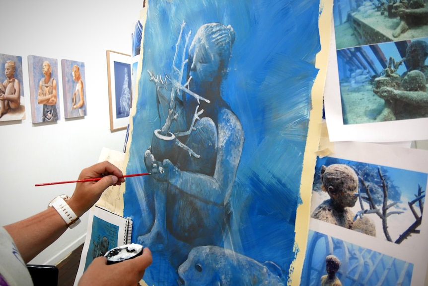 A woman paints a picture of an underwater sculpture with reference photos