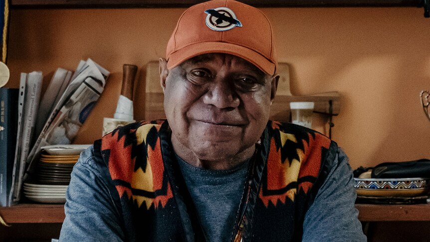 Archie Roach wears a cap and vest and looks into the camera smiling