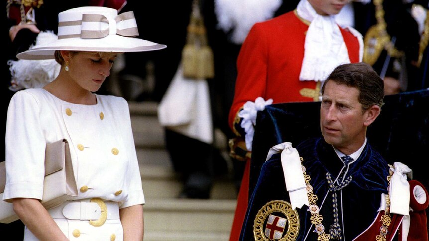 Princess Diana stands next to Prince Charles who is sitting down while someone in a red coat holds the Prince's coat-tails.
