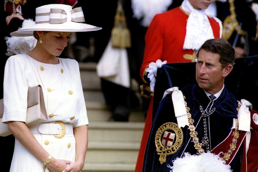 Princess Diana stands next to Prince Charles who is sitting down while someone in a red coat holds the Prince's coat-tails.