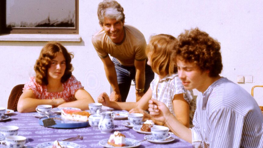 Colour 1980s photo of family sitting around a table, with plates of cake in front of them.