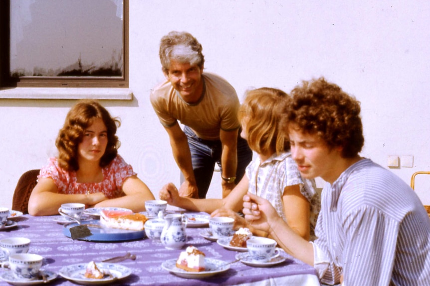 Colour 1980s photo of family sitting around a table, with plates of cake in front of them.