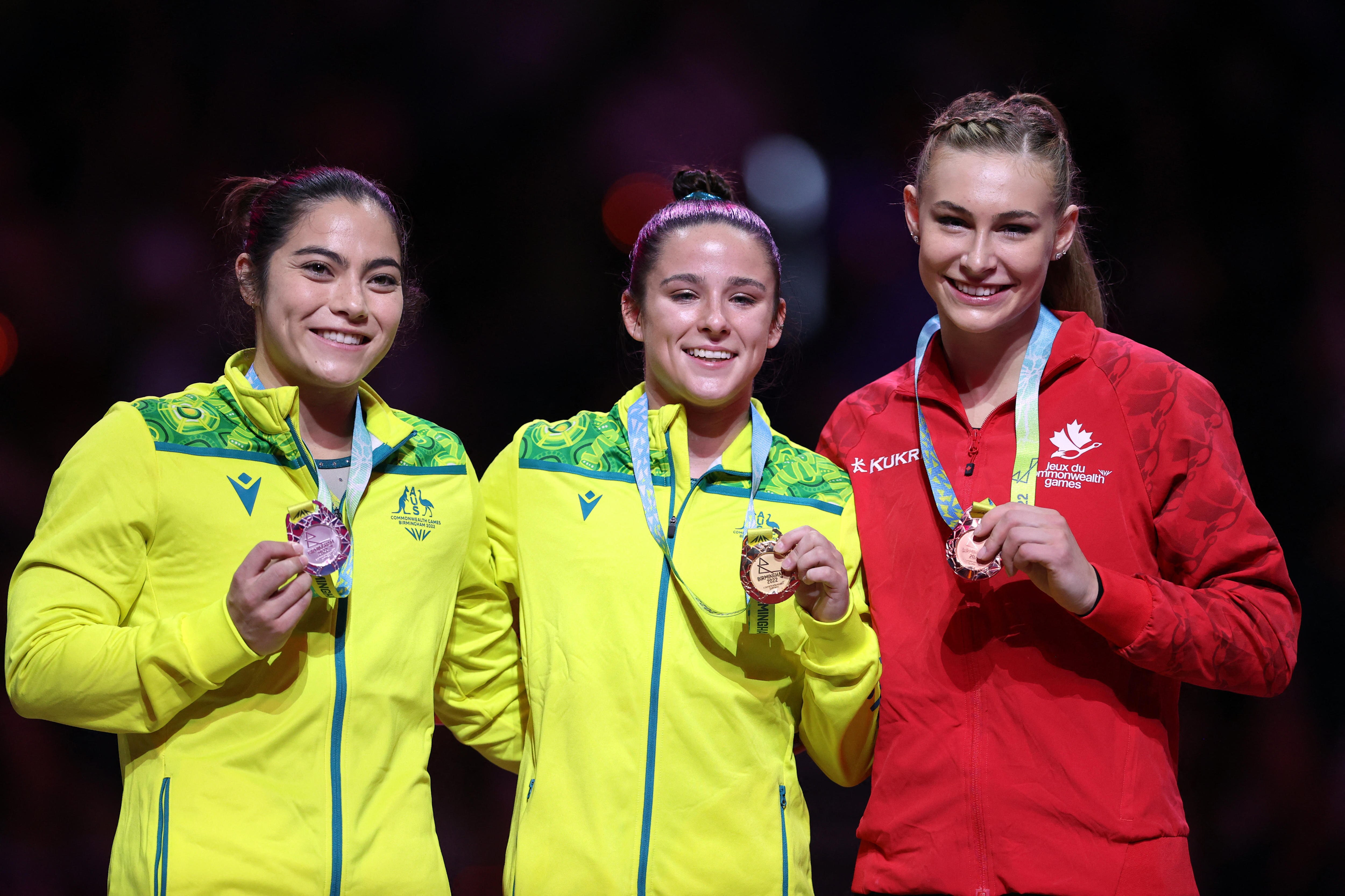 Two smiling Australian gymnasts stand on the podium with their medals along with a Canadian competitor. 