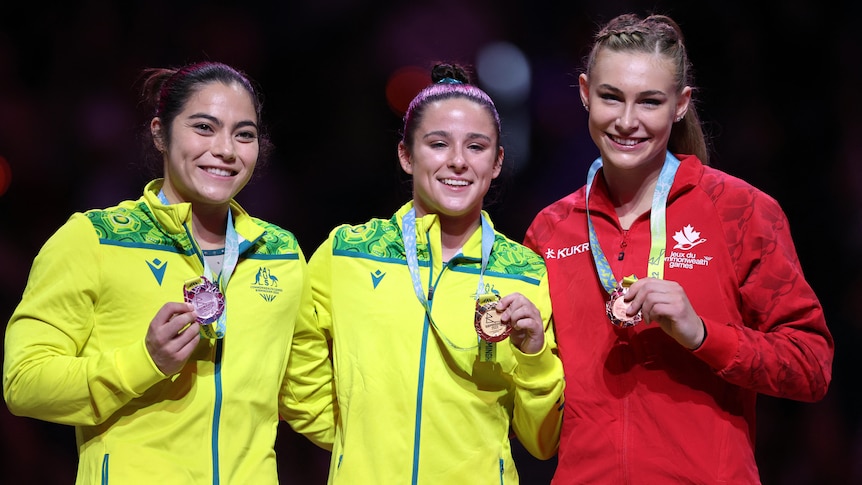 Two smiling Australian gymnasts stand on the podium with their medals along with a Canadian competitor. 