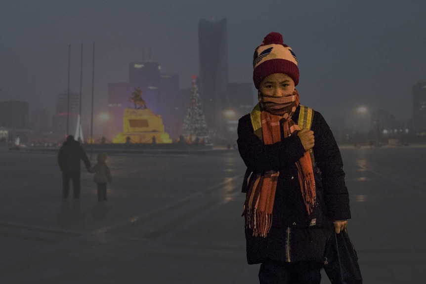 A child, rugged up with a scarf over their mouth, stands in front of a dark, smoggy city.