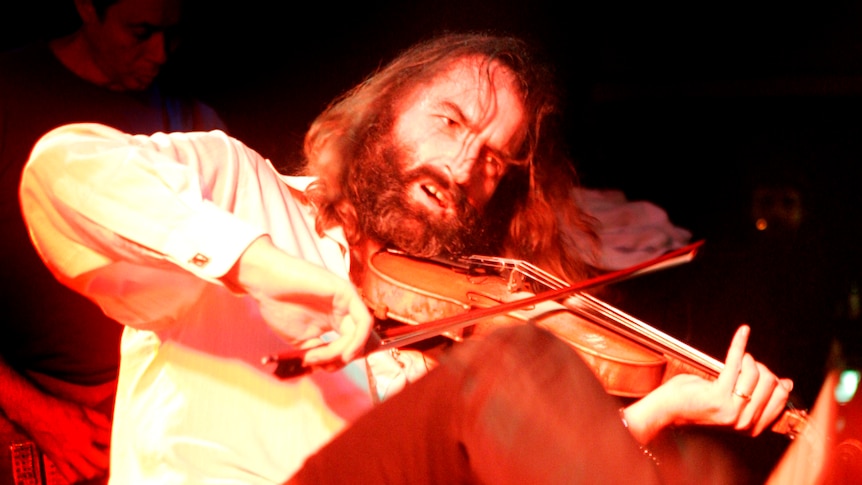 Warren Ellis plays violin and kicks his leg up. He wears a white shirt, a bushy beard and long hair.